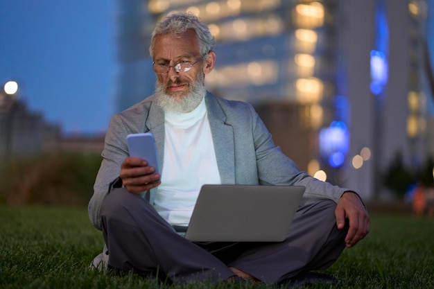 Old business man sitting outside office in city park using\
smartphone at night