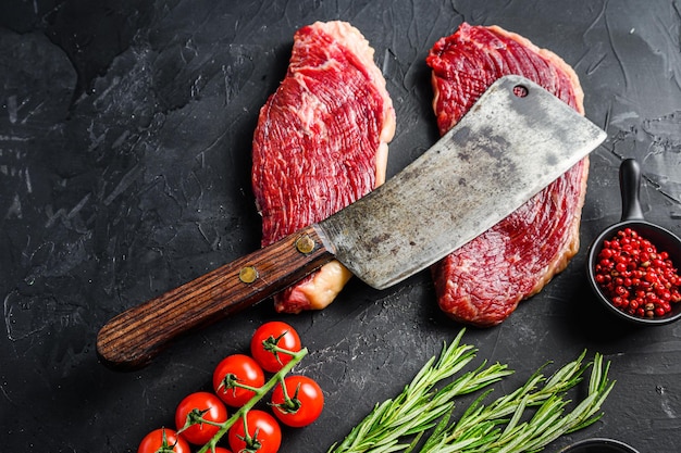 Old butcher cleave over raw picanha beef steaks with rosemary, peppercorns and salt on black textured background, top view Close up.