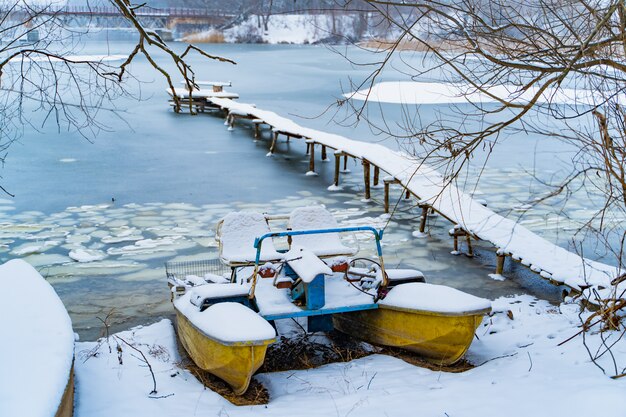 Old catamaran in the winter on the snow coast of the frozen river next to the long bridge. 