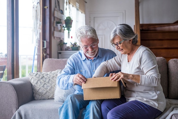 Old couple unpacking delivery box at home Happy senior man and woman looking at carton box while sitting on sofa in living room Elderly husband and wife checking out delivered stuff on couch
