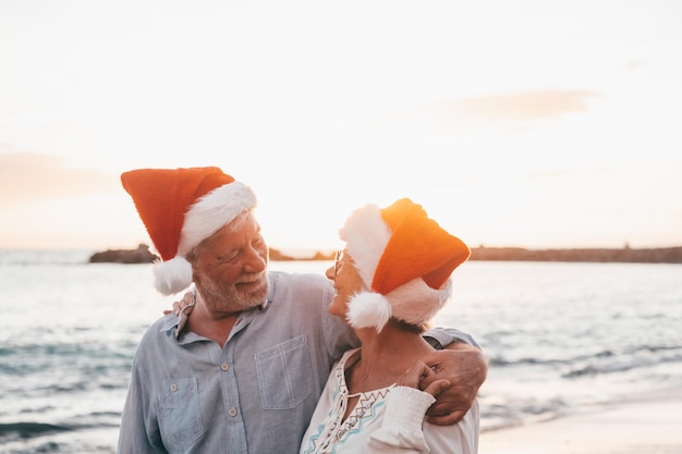 Old cute couple of mature persons enjoying and having fun together at the beach wearing christmas hats on holiday days Hugged on the beach with the sunset at the background at winterxA