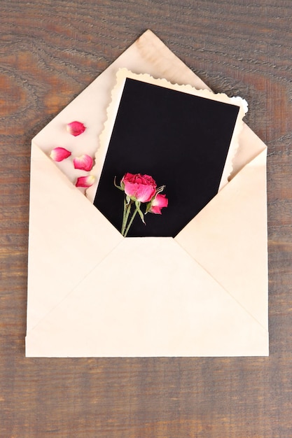 Old envelope with blank photo paper and beautiful pink dried roses on wooden background