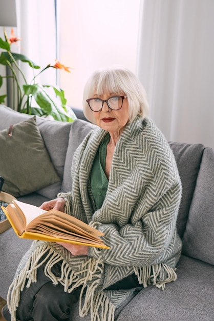 Vecchia donna anziana seduta sul divano a leggere un libro a casa