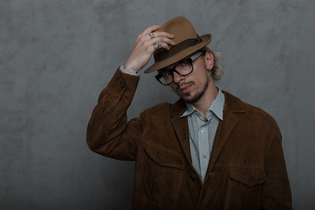 Old-fashioned young man hipster with a beard straightens a stylish hat in a room near a gray wall.