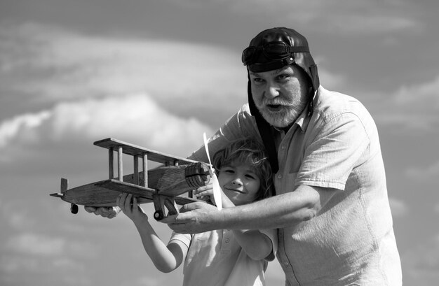 古い祖父と幼い子供の孫が夏の空の背景に木製の飛行機で遊んでいる