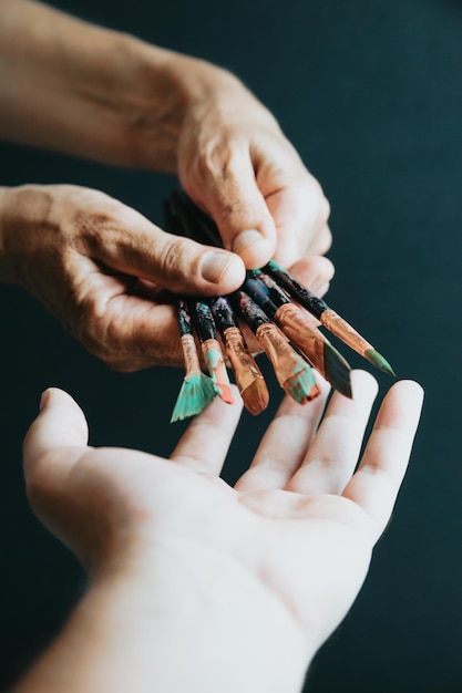 Old hands giving painting pencils brushes to another hand, black background, moody style. Artist style. art concept