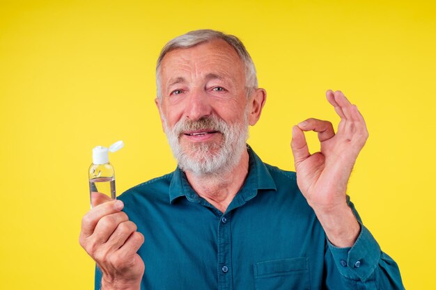 Old happy senior man wearing green cotton shirt with a collar and applying alcohol gel to make cleaning and clear germ, bacteria yellow background.