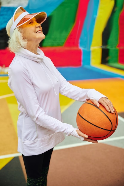 Signora anziana, sana e allegra con la palla da basket su una piazza colorata all'aperto