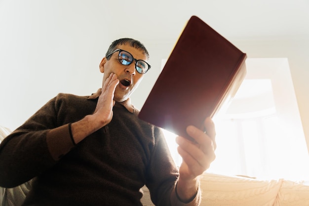 Old indian man reading a book at home wearing glasses scared in shock with a surprise face afraid an