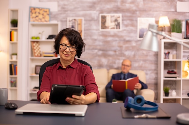 Old lady in her 60s using modern technolgy in cozy living space while her husband reads in the background