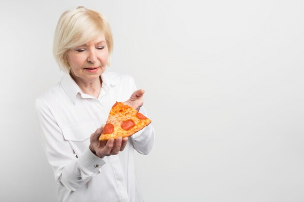 Old lady holding a big piece of pizza in her hands. She is trying to guess what ingredients are in it. Also she is thinking about eating that piece.