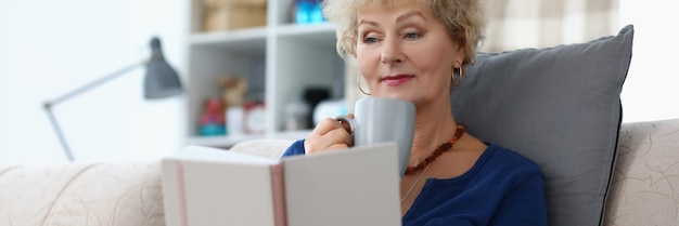 Old lady reading book and drinking cup of coffee being comfy on sofa at home