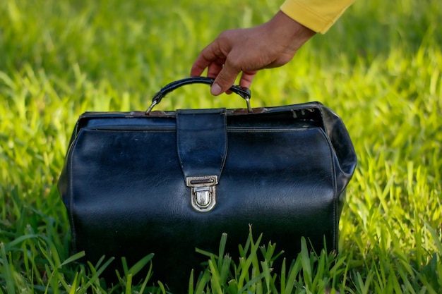 Старый кожаный чемодан, который держится рукой на зеленой траве, сверху у него пустая карточка.