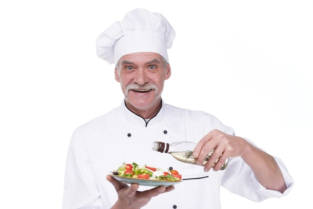 Old male cook holding a plate with vegetables salad and oil on white wall