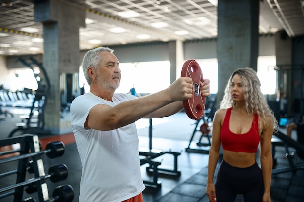 Old man doing exercise with bar, female personal trainer, gym interior on background. Sportive grandpa with woman instructor, workout in sport center