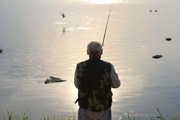 Foto vecchio che pesca pescatore anziano dai capelli grigi lancia una rotazione dalla riva al tramonto girando una bobina maschio anziano positivo che pesca al lago rotando la canna rotante pescatore in pensione che si gode il fine settimana