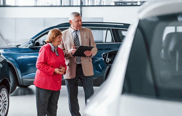 Old man in formal wear with notepad supporting woman in choosing automobile.