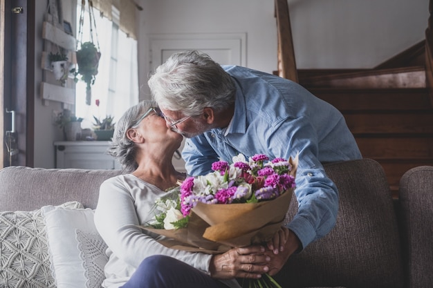 サンバレンタインデーのために自宅のソファに座っている彼の妻に花を与える老人。一緒に驚きのキスを楽しんでいる年金受給者。愛の人々が楽しんでいます。