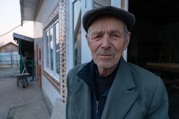 old man in grey blazer and hat stands near his house