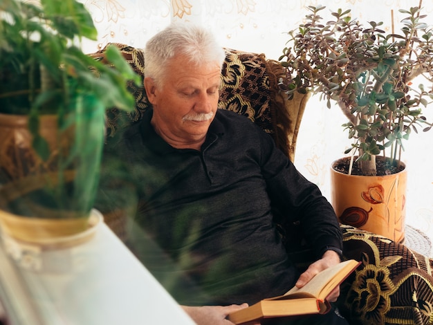 Old man is reading a book while sitting in an armchair.