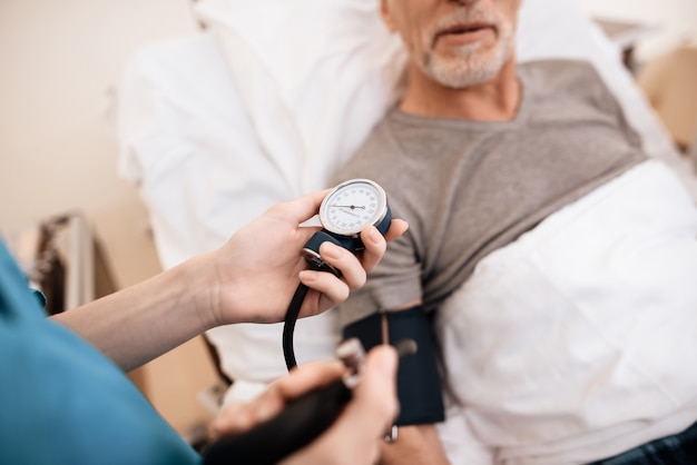 Old man lies on a cot in the ward, nurse measures pressure.