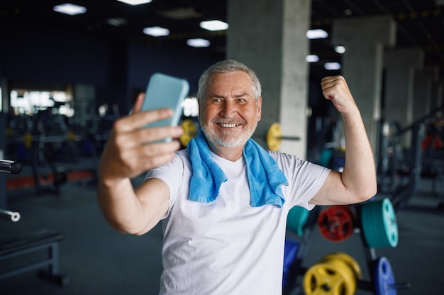 Old man shows his muscles and makes selfie, gym