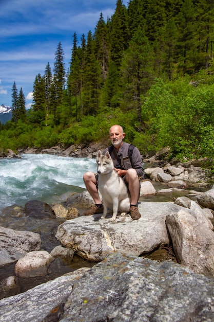 老人とそり犬は川の近くを歩きます。高山の風景です。アクティブなレジャー年金受給者。老人は笑っています。シベリアンハスキーと一緒に歩きます。