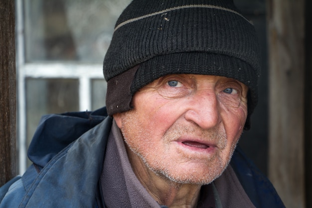 old man in sloppy clothes stands on the threshold of his own ruined house and looks into the distance