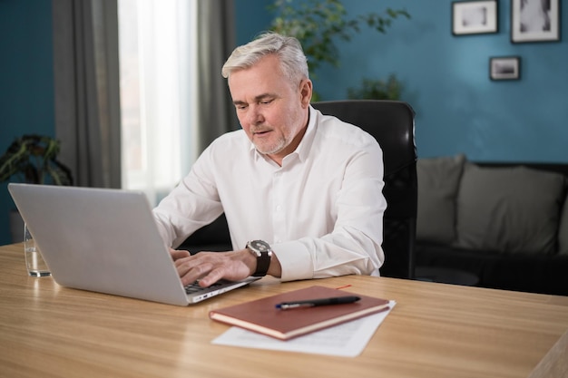 Old man using laptop smiling at living room at home working in quarantine