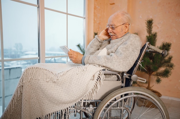 Старик в инвалидной коляске сидит у большого окна и пользуется планшетом
