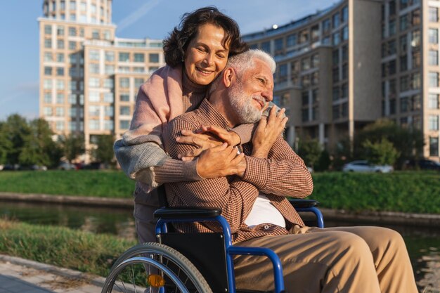 Old man in wheelchair walking with caregiver senior woman on road in park elderly family couple