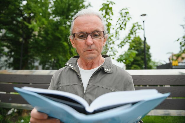 Старик с седыми волосами читает книгу на скамейке в парке Отдых в парке