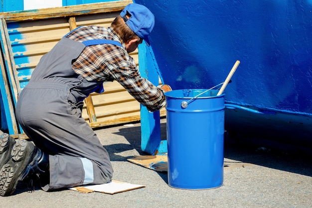 Un vecchio con una tuta da lavoro dipinge un bidone della spazzatura o un contenitore per strada con un pennello