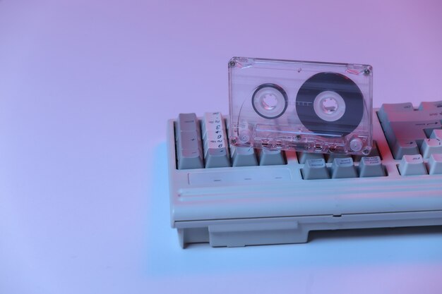 Old pc keyboard with audio cassette in pink blue gradient neon, holographic light. Retro Attributes 80s