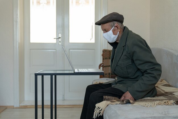 Uomo anziano pensionato in maschera medica viso protettivo seduto sul divano di casa vicino al tavolo da lavoro con il computer portatile