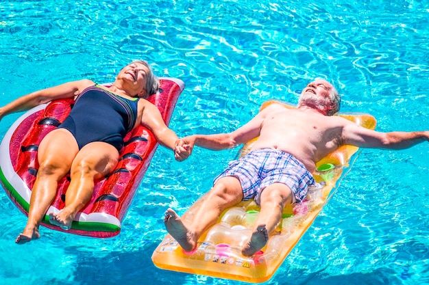 Le coppie anziane di anziani si rilassano e dormono sull'acqua limpida della piscina blu si sdraiano su materassini gonfiabili colorati alla moda e si prendono per mano con amore per il concetto di stile di vita per sempre insieme