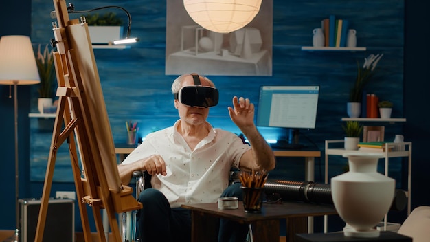 Old person with disability in wheelchair using vr glasses to draw professional artwork masterpiece on canvas. Suffering from chronic impairment, working with 3d augmented reality.