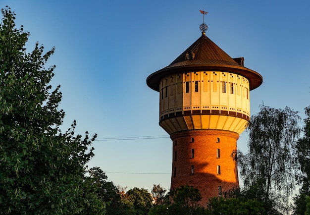 Старая водонапорная башня из красного кирпича, окруженная деревьями в свете заката