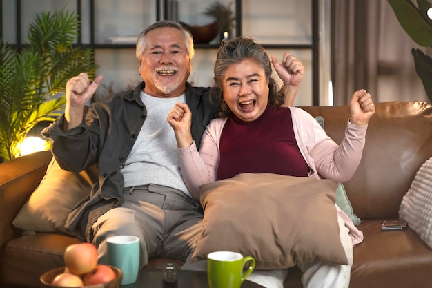 ホームオールド成熟したアジアのカップルでテレビを見ている古い引退した年齢のアジアのカップルは、リビングルームの家の隔離活動でソファのソファで笑い笑顔の勝利と一緒にスポーツゲームの競争を応援しています