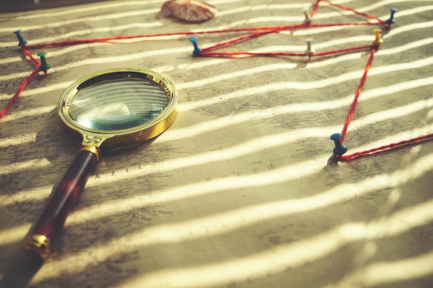 Old retro map is pressed with magnifying glass, route marked on
map with red thread and buttons.sunlight shines through the
blinds