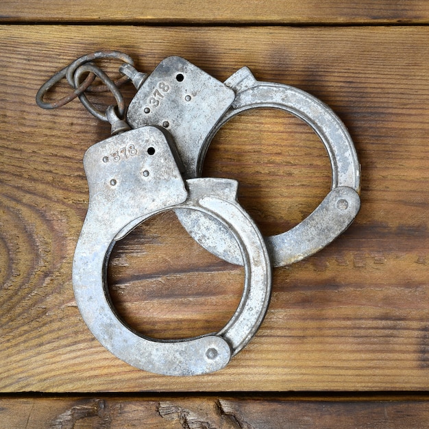 Old and rusty police handcuffs lie on a scratched wooden surface. 