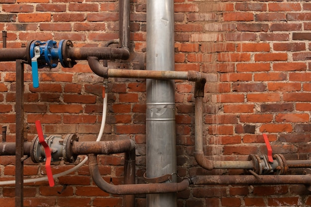 Old rusty water pipes with valves in an industrial building opposite a brick wall.