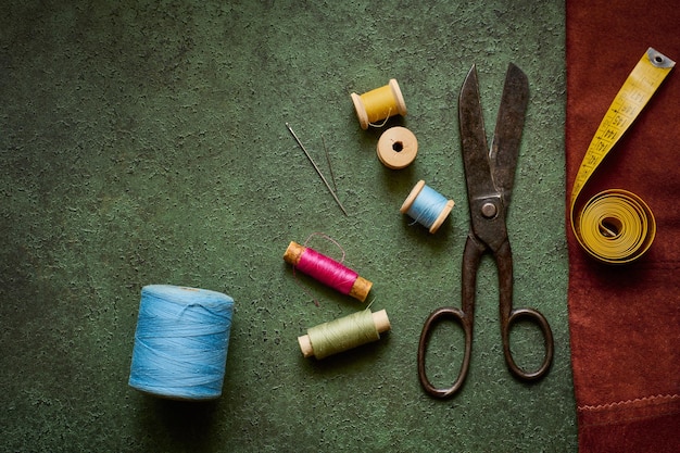 Old scissors, tailor tape and spools of thread on a dark green\
textured background close up, flat lay