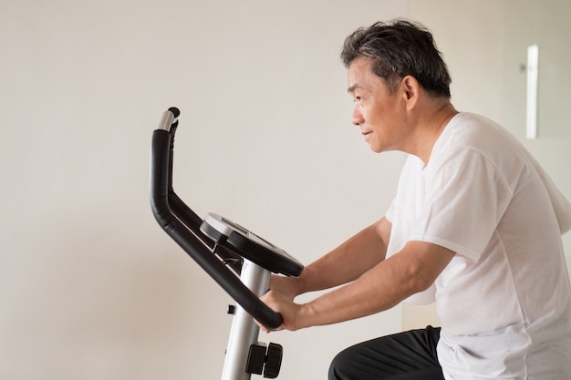 Il vecchio uomo anziano in bicicletta, esercizio, allenamento in palestra con la moderna macchina da ciclismo