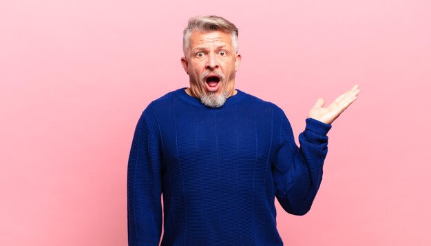 Old senior man looking surprised and shocked, with jaw dropped holding an object with an open hand on the side