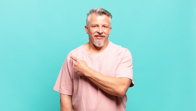 Old senior man smiling cheerfully, feeling happy and pointing to the side and upwards, showing object in copy space