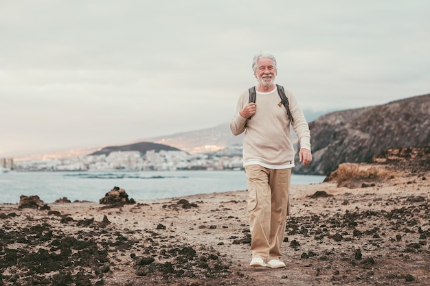 Uomo anziano anziano che cammina sulla scogliera indossando zaino e sorridente, godendosi un'escursione in mare alla luce del tramonto. pensionato dai capelli bianchi attivo