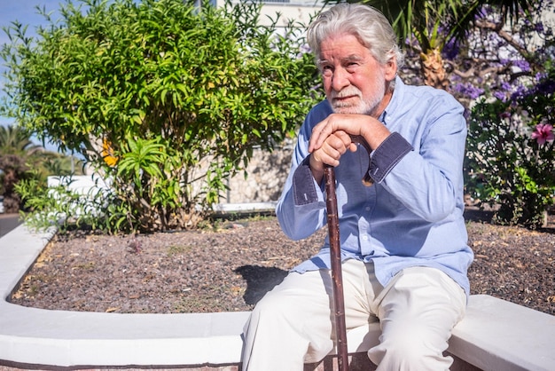 Old senior man with hands over the cane sitting in public park with sad expression Elderly retiree with back pain using walking stick Tree and flowers on background