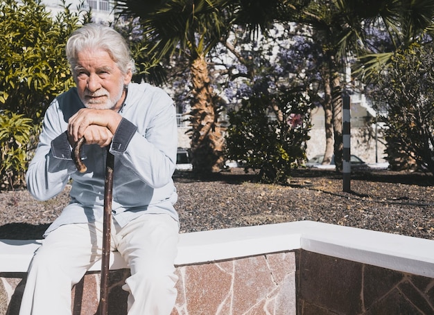 Old senior man with hands over the walking cane sitting in public park with sad expression