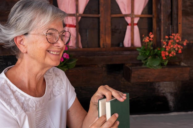 Старая пожилая женщина в белом сидит на улице дома и держит книгу, улыбаясь